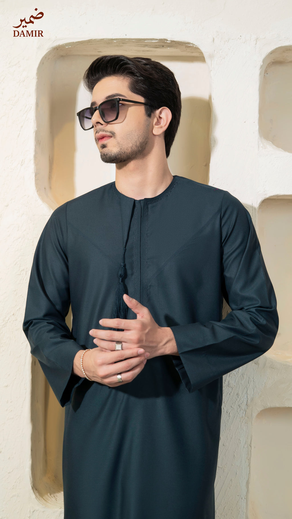 Man wearing a Teal traditional outfit with sunglasses, standing against a light-colored wall.