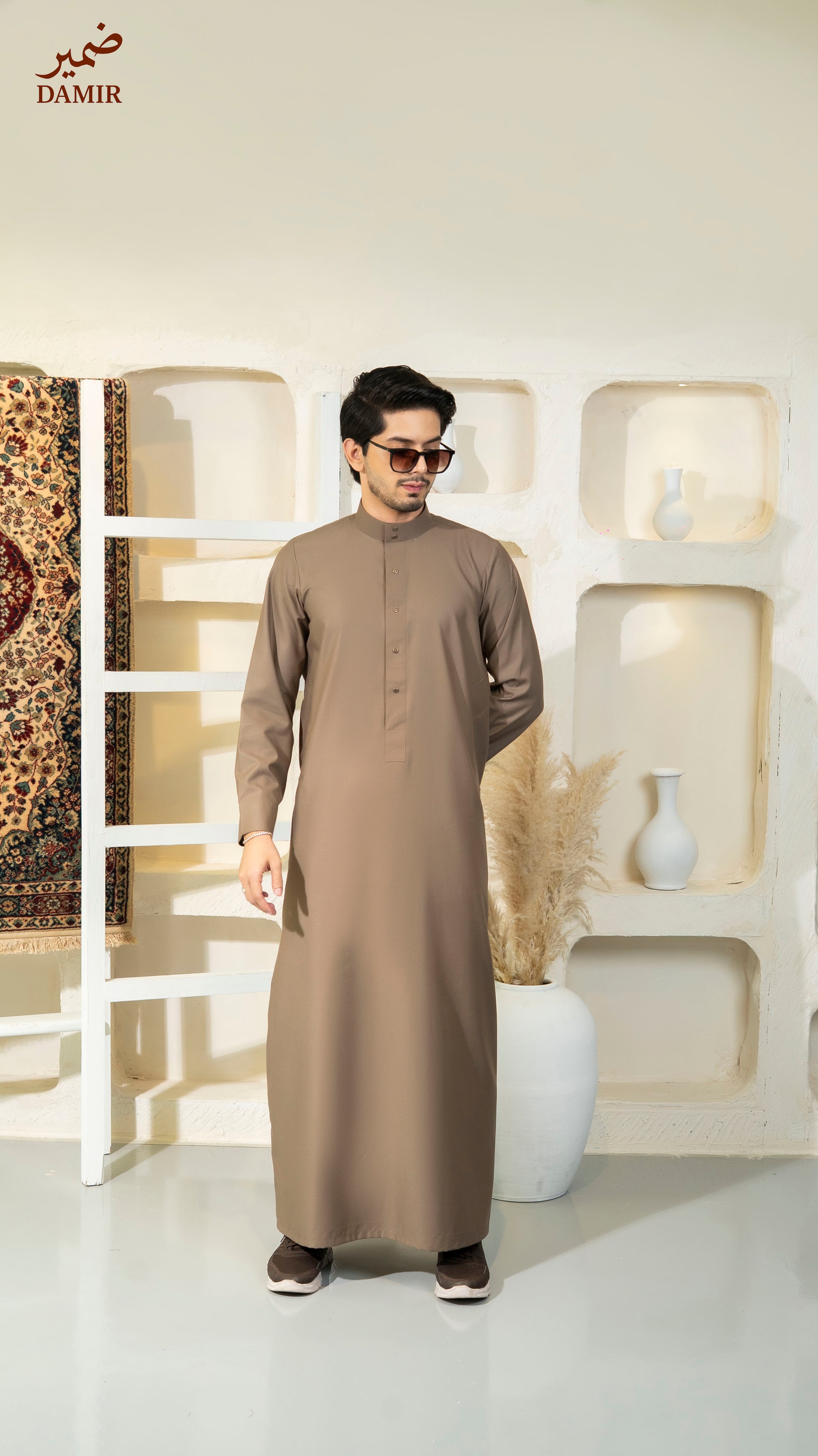 Man wearing a brown traditional outfit standing in a modern interior setting with 'DAMIR' branding.