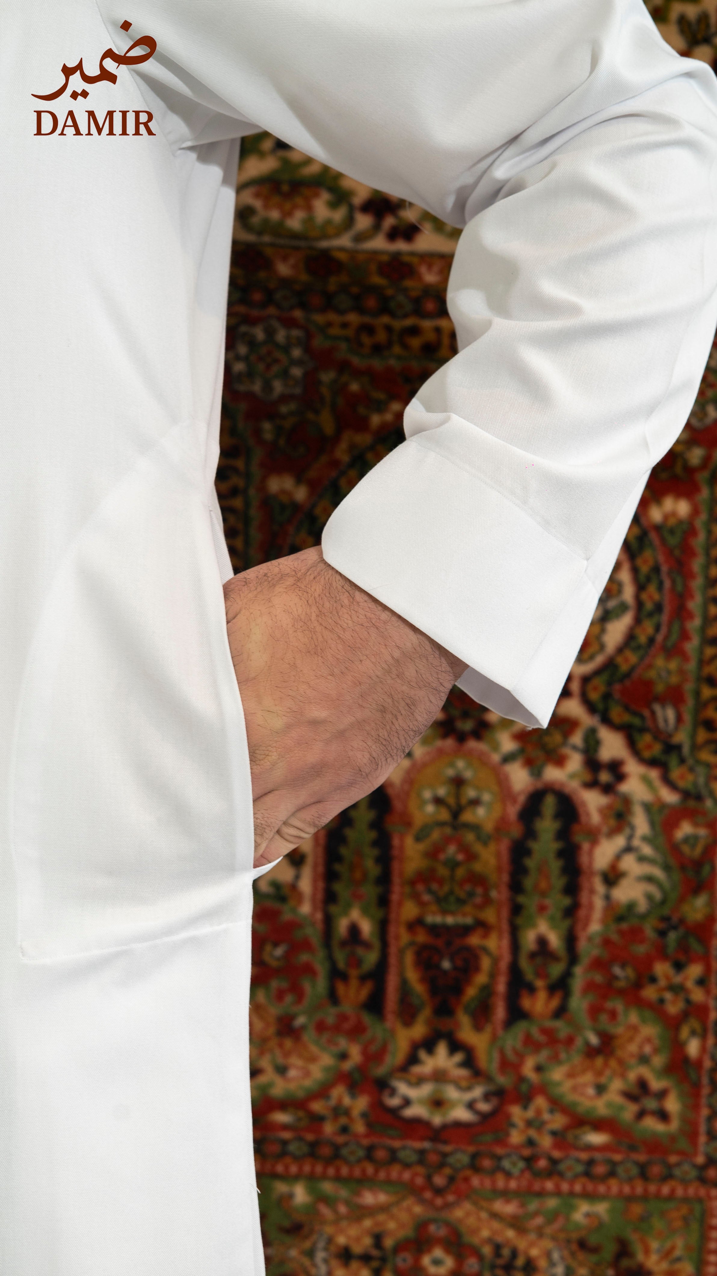 Close-up of a white garment with a patterned rug in the background, featuring the brand 'DAMIR'.